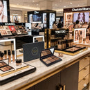 An open Flouréa Beauty Shop 6-pan eyeshadow palette and its dark grey branded box are positioned centrally on a marble counter in a brightly lit luxury department store. The product is surrounded by large, illuminated retail displays for major competitor brands including Chanel, Tom Ford, Dior, and Charlotte Tilbury.