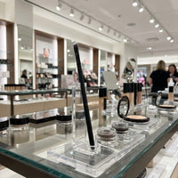 A close-up view of the Flourea Angled Liner Brush standing on a glass retail counter next to open makeup testers, set against the blurred backdrop of a busy, upscale beauty department store aisle.