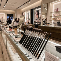 The Flourea Angled Liner Brush displayed upright in a sleek black holder on an illuminated glass counter inside a luxury department store beauty section, surrounded by mirrors and other cosmetic displays.