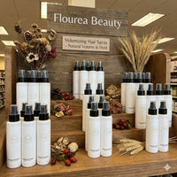 Wooden retail merchandising display stocked with multiple bottles of Flourea Volumizing Hair Spray, decorated with dried florals and wheat stalks. Signage reads "Flourea Beauty" and "Natural Volume & Hold."