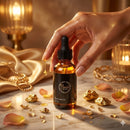 Hand holding a bottle of Flourea's Rose Gold Oil with gold flakes and rose petals on a marble surface.