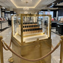 A locked, high-security brass and glass display case housing the full Flourea Foundation collection on marble tiers, surrounded by velvet rope stanchions on the marble floor of a luxury department store beauty hall.