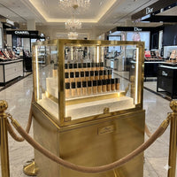 A locked, high-security brass and glass display case housing the full Flourea Foundation collection on marble tiers, surrounded by velvet rope stanchions on the marble floor of a luxury department store beauty hall.